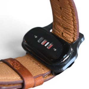 May include: Close-up of a black device attached to a brown leather watch band. The device has a glossy finish with small, illuminated indicators in red and white. The watch band is a light brown color with visible stitching.