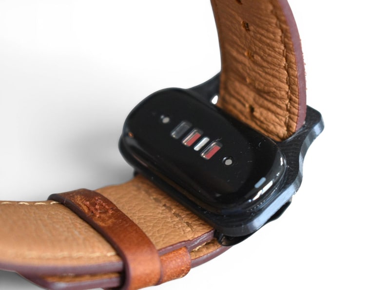 May include: Close-up of a black device attached to a brown leather watch band. The device has a glossy finish with small, illuminated indicators in red and white. The watch band is a light brown color with visible stitching.