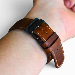 May include: Close-up of a brown leather watch strap with white stitching and a black buckle. The strap is secured on a wrist, showcasing the details of the leather and the stitching.