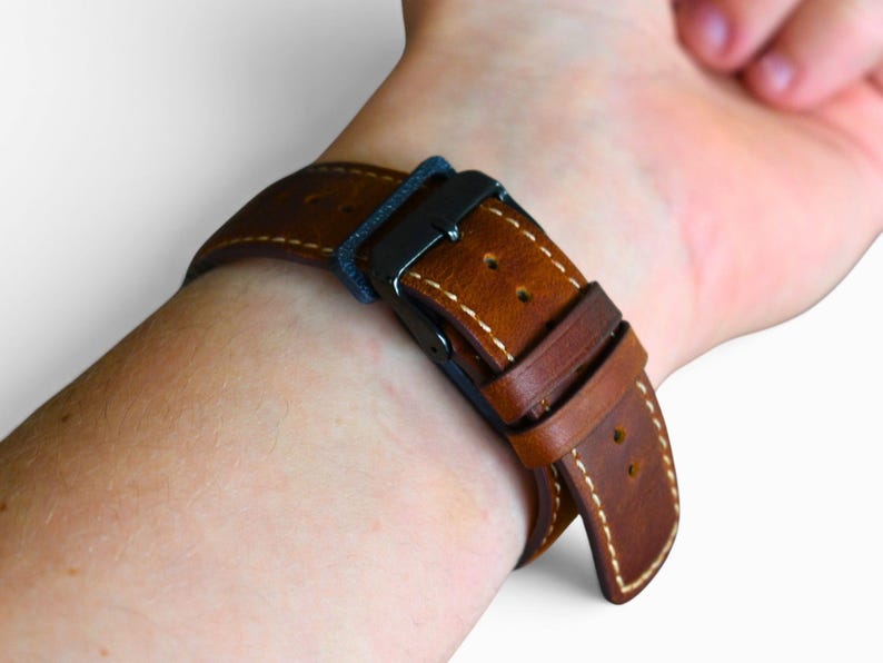 May include: Close-up of a brown leather watch strap with white stitching and a black buckle. The strap is secured on a wrist, showcasing the details of the leather and the stitching.