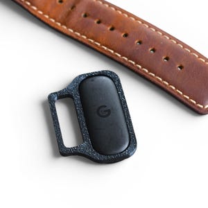 May include: A black rectangular device with a small handle, featuring a textured frame and a centered 'G' logo, sits near a brown leather watch strap with white stitching. The strap is a warm brown color.