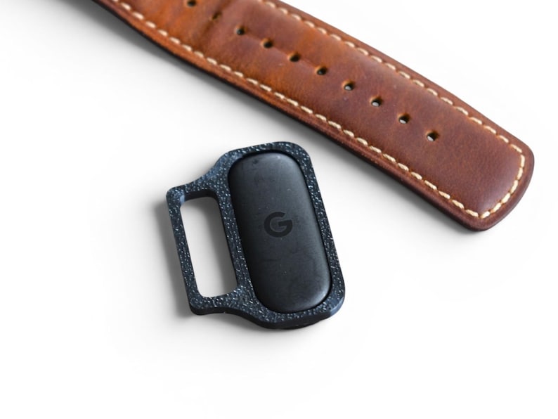 May include: A black rectangular device with a small handle, featuring a textured frame and a centered 'G' logo, sits near a brown leather watch strap with white stitching. The strap is a warm brown color.