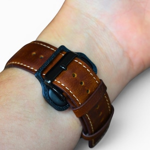 May include: A brown leather watch band with white stitching and a black buckle is fastened on a wrist. The watch band is made of a rich, textured leather. The watch band is secured with a black buckle.