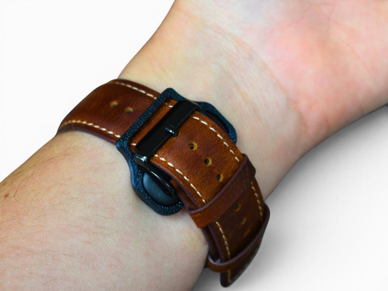 May include: A brown leather watch band with white stitching and a black buckle is fastened on a wrist. The watch band is made of a rich, textured leather. The watch band is secured with a black buckle.