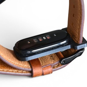 May include: Close-up of a brown leather watch band with a black rectangular device attached. The band is secured with a black metal buckle. The device has a black glossy finish and a series of small red lights.