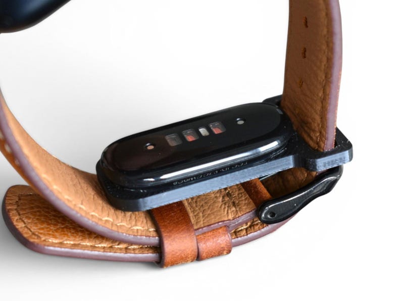 May include: Close-up of a brown leather watch band with a black rectangular device attached. The band is secured with a black metal buckle. The device has a black glossy finish and a series of small red lights.