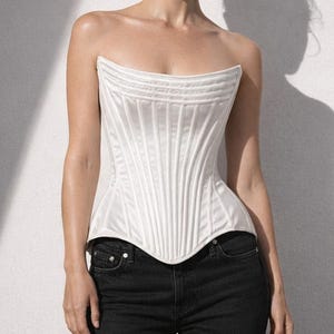 Op de afbeelding: Een witte, strapless corset met verticale panelen en een gebogen halslijn. Het corset wordt gecombineerd met een zwarte denim jeans. De afbeelding is geplaatst tegen een effen witte muur, met een schaduw op de muur.