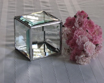 May include: A clear glass cube-shaped box with silver edging. A bouquet of pink roses sits beside the box. The glass reflects light, creating a prism effect. The box could be used for jewellery or small keepsakes.