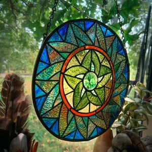 May include: A stained glass suncatcher with a circular design featuring a central green flower with a diamond-shaped jewel in the centre. The flower is surrounded by a red ring and a blue and green outer ring.