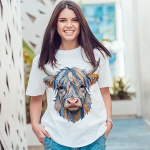 May include: A white t-shirt with a colourful patchwork design of a Highland cow's face. The cow's fur is rendered in blue, pink, and yellow hues, with curved horns and a pink nose. The shirt has a crew neck and short sleeves.