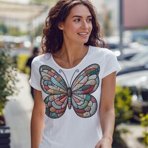 以下が含まれることがあります： カラフルな蝶のデザインが施された白いTシャツ。蝶は、赤、青、緑、ピンクの色合いの様々な幾何学的な形で構成されています。このシャツは半袖で、クルーネックです。