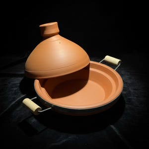 May include: A terracotta tagine pot with a conical lid, partially open. The pot has a round base with a metal rim and wooden handles. The lid features a knob and decorative lines. The color is a warm, earthy orange.