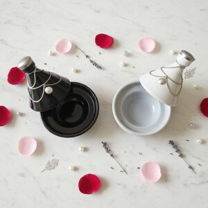 Peut inclure: Deux récipients décoratifs en céramique, un noir et un blanc, avec des couvercles argentés et des détails de chaîne. Les récipients sont entourés de pétales de rose, de perles et de brins de lavande sur une surface en marbre.