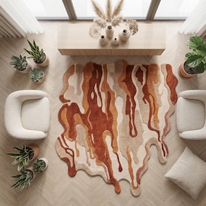 May include: An overhead view of a modern living room with a unique rug. The rug features a flowing, abstract design in shades of brown, beige, and cream. Two cream-colored armchairs and several potted plants surround the rug, creating a cozy atmosphere.