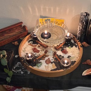 May include: A circular wooden tray containing a glass bowl filled with water and a floating black candle. The tray is decorated with white wax, cinnamon sticks, star anise, seashells, and four small black candles. Nearby are incense, candles, and a plant.