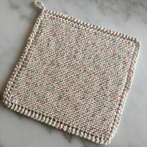 Pode incluir: Um pegador de panela quadrado, de malha branca suja, com uma alça para pendurar. É salpicado com pequenas manchas rosa, azul e verde. As bordas são finalizadas com uma borda decorativa.