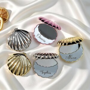 May include: A collection of shell-shaped compact mirrors in gold, silver, and rose gold finishes. Three mirrors are open, revealing reflective surfaces with the names Omaira, Alice, and Sophia engraved. Two dried citrus slices are visible nearby.