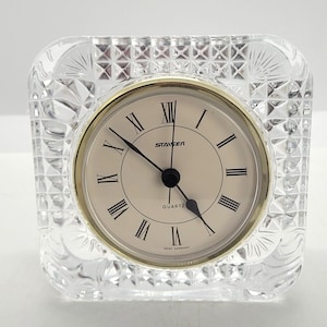 May include: A clear, square quartz clock with a gold-colored bezel and Roman numeral hour markers. The clock face is cream-colored, and the clock is encased in a cut-glass design. The clock face reads "STAVGER QUARTZ WEST GERMANY".