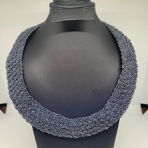 May include: A dark grey beaded necklace on a black mannequin. The necklace is thick and woven, with a textured appearance. The beads are small and evenly spaced, creating a solid, substantial piece of jewellery.
