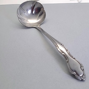May include: A silver-colored ladle with a rounded bowl and a long, decorative handle. The bowl has a petal-like design. The handle features ornate detailing. The ladle is made of a shiny, reflective metal.