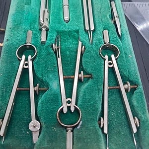 May include: A set of vintage drafting tools, including several compasses and other instruments, neatly arranged in a green velvet-lined case. The tools are made of silver-colored metal and appear to be well-preserved, suggesting careful use and storage.