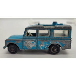 May include: A vintage die-cast toy Land Rover in blue and gray, showing signs of wear. The toy car features a detailed design with visible windows, wheels, and a roof rack. The paint is chipped, giving it a classic, aged appearance.