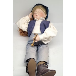 May include: A porcelain doll depicting a young boy in a blue waistcoat, white shirt, and grey trousers. He wears a dark blue hat and brown boots. The doll holds a gold-coloured trumpet and appears to be asleep. A small woven basket is behind the doll.