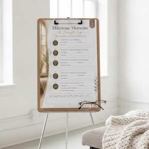 Op de afbeelding: Een klembord op een statief met een Milestone Moments & Strength Log-blad met vijf genummerde fitnessdoelen, waaronder pull-up mijlpalen. Het papier heeft gouden en bruine accenten en er ligt een bril met bruin montuur op het blad.