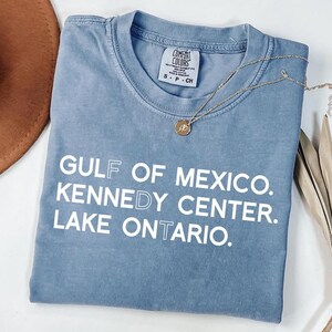 Op de afbeelding: Een blauw t-shirt met korte mouwen, een ronde hals en een Comfort Colors merklabel. Het shirt is voorzien van witte tekst met de woorden GULF OF MEXICO, KENNEDY CENTER en LAKE ONTARIO, waarbij specifieke letters een lichtblauwe omlijning hebben.