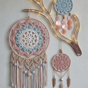 May include: Three circular dream catchers with crochet mandalas in shades of pink, blue, and cream. The largest features macrame fringe with beads. A light blue dream catcher has shell discs, and a small pink one has gold-toned feather charms.