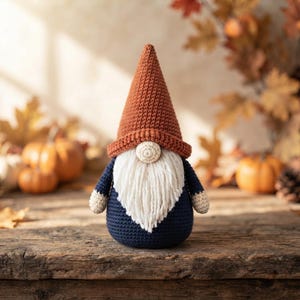 Peut inclure: Une figurine de gnome au crochet avec un grand chapeau pointu orange rouille, un nez crème, une longue barbe en fil blanc et un corps bleu marine foncé avec des manches crème. Le gnome présente un motif de points texturé et visible.