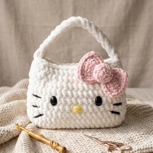 Può includere: Una borsa bianca all'uncinetto con un fiocco rosa e dettagli neri. La borsa ha un manico ed è appoggiata su una superficie in maglia color crema. Un uncinetto e delle forbici sono in primo piano.