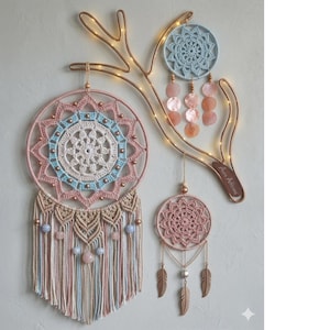 May include: A decorative wall hanging featuring three dream catchers in shades of pink, blue, and cream. The largest dream catcher has a detailed crochet design and beaded accents. A copper-colored metal antler shape holds the dream catchers, with small lights adding a warm glow.