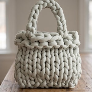 May include: A light sage green handbag crafted from thick, chunky knit yarn. The bag features a structured, rectangular body with a wide, braided top opening and two integrated, braided handles that extend from the top edge of the bag.