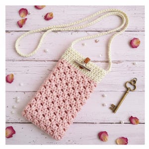 May include: A handmade crochet phone crossbody bag in pink and cream. The bag features a woven pattern, a cream-colored strap, and a silver-tone clasp with a leather accent. The bag is shown on a light wood surface with decorative elements.