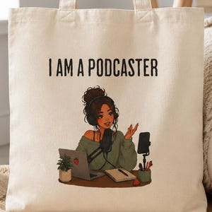 May include: Cream-coloured tote bag featuring the words "I AM A PODCASTER" and an illustration of a woman podcasting. The illustration shows a woman with a microphone, laptop, and a smartphone. Black headphones and a notebook are on the table.