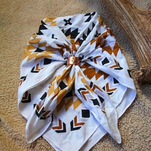 May include: White bandana with a geometric pattern in black and gold, secured with a copper ring. The bandana features a repeating design of arrows, triangles, and crosses. The copper ring has an engraved design.