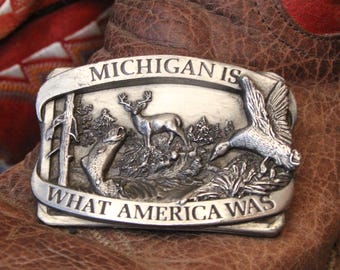 May include: A rectangular silver-toned metal belt buckle featuring a relief scene of a deer, a fish, and a flying duck. The text MICHIGAN IS WHAT AMERICA WAS is embossed on the top and bottom banners of the buckle, which rests on a brown boot.