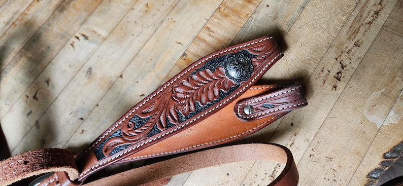 May include: Close-up of a brown leather belt with intricate floral tooling and a silver-toned buckle. The belt features a detailed design with dark accents and white stitching, showcasing a rustic, handcrafted aesthetic.