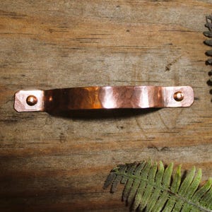 May include: A hammered copper drawer pull with a curved, arched handle design. The hardware features flat, rectangular ends, each secured with a single small, rounded copper rivet. The metallic surface has a polished, reflective finish.