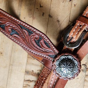 May include: Close-up of a brown leather horse bridle with intricate floral tooling and silver-colored hardware. The bridle features a decorative silver concho and a detailed, carved design with black accents.