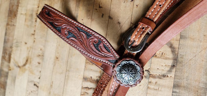 May include: Close-up of a brown leather horse bridle with intricate floral tooling and silver-colored hardware. The bridle features a decorative silver concho and a detailed, carved design with black accents.