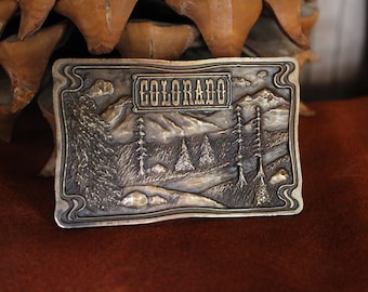 May include: A rectangular metal belt buckle with a raised relief design featuring a mountain landscape, pine trees, and a river. The word COLORADO is embossed in a rectangular frame at the top center. The buckle has a metallic finish with dark patina.