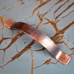 May include: A copper-colored cabinet pull with a curved handle. The rectangular ends have pre-drilled holes for mounting. The hardware has a distressed finish, suggesting an antique or rustic style. The pull is shown against a weathered, gray-blue surface.