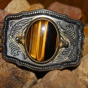 May include: A rectangular belt buckle featuring a polished silver-tone metal frame with intricate scrollwork engravings and a beaded border. A large, oval-shaped tiger's eye gemstone with distinct golden-brown and black chatoyant bands is centered.