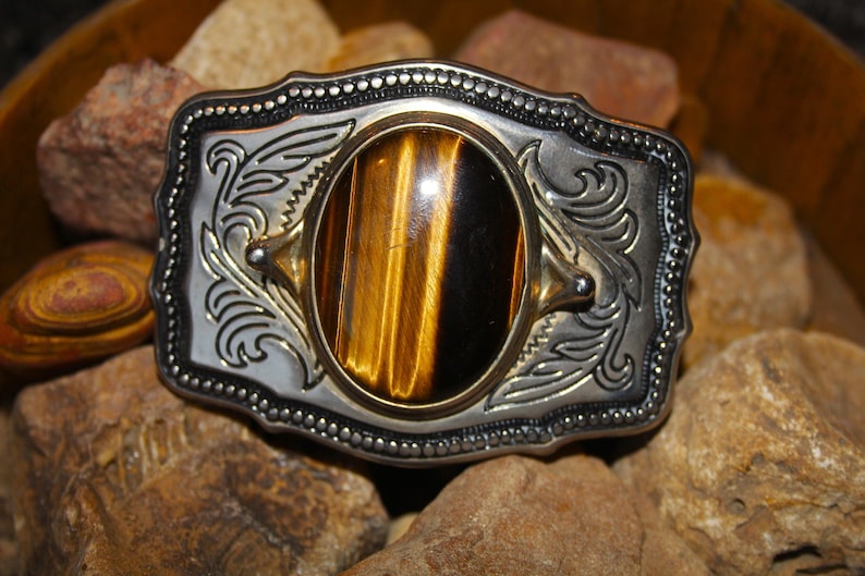 May include: A rectangular belt buckle featuring a polished silver-tone metal frame with intricate scrollwork engravings and a beaded border. A large, oval-shaped tiger's eye gemstone with distinct golden-brown and black chatoyant bands is centered.