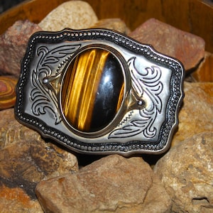 May include: A rectangular silver-tone metal belt buckle featuring an oval-shaped tiger's eye gemstone at the center. The metal surface is decorated with intricate engraved scrollwork patterns and a beaded border along the outer edge.