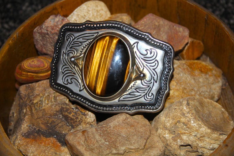 May include: A rectangular silver-tone metal belt buckle featuring an oval-shaped tiger's eye gemstone at the center. The metal surface is decorated with intricate engraved scrollwork patterns and a beaded border along the outer edge.