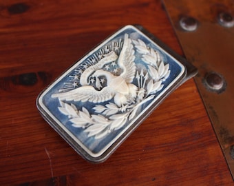 Puede incluir: Un objeto decorativo rectangular azul y blanco con un relieve detallado de un águila con las alas extendidas, estrellas y ramas de laurel. El objeto tiene un marco de metal plateado y descansa sobre una superficie de madera.