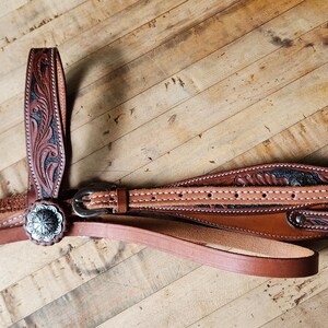 May include: A brown leather horse bridle with intricate floral tooling and a silver-colored concho. The bridle features multiple straps, a buckle, and a browband. The background is a wooden surface.